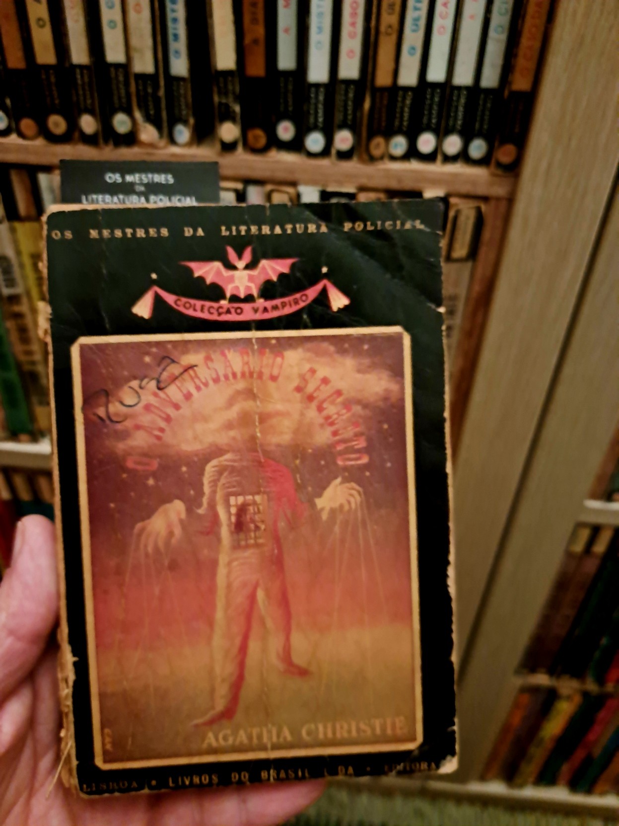 A picture of Agatha Christie's "The Secret Adversary" in its Portuguese edition on Colecção Vampiro. The series' tag line "Os mestres da literatura policial" can be read on the book cover and also the bookmark on it. On the background, a bookshelf full of books of the same collection.