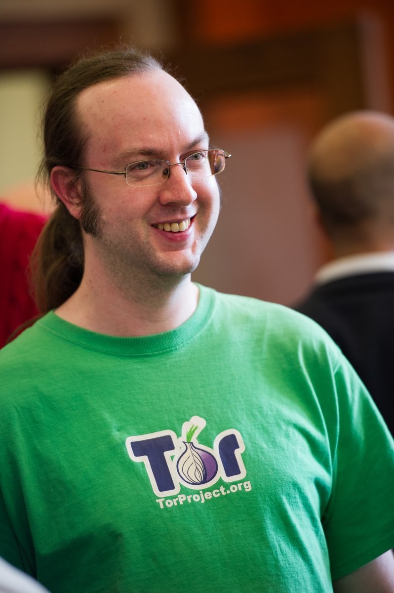 Um homem de óculos e rabo de cavalo a sorrir. Tem uma t-shirt verde claro com o logótipo do Tor.