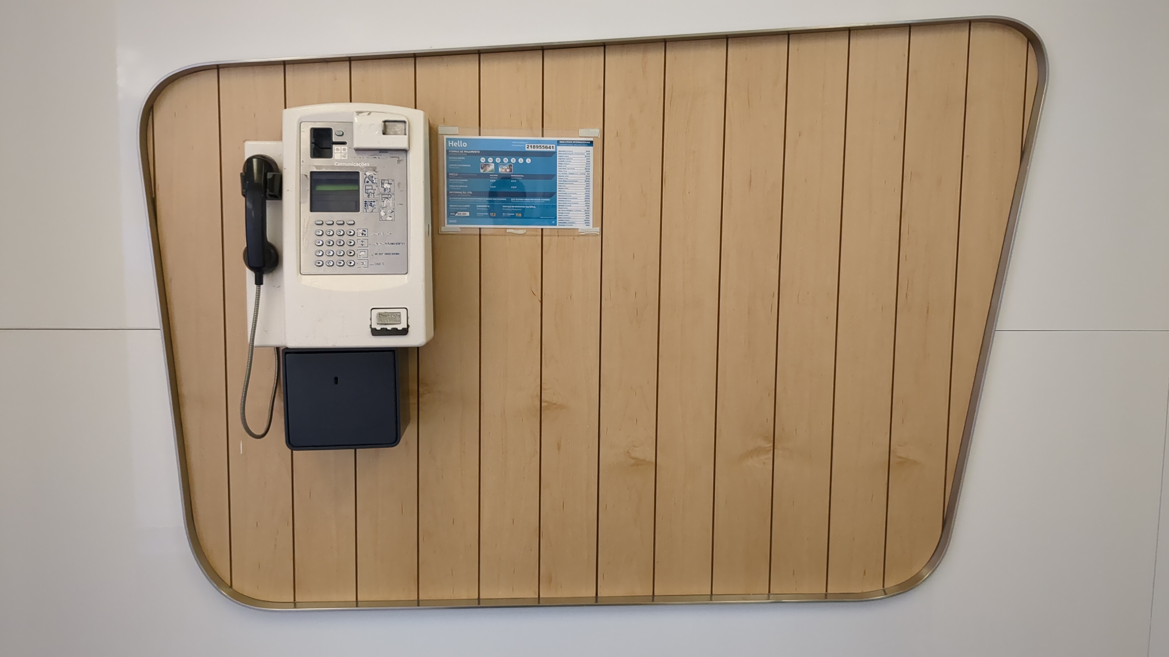 Telefone público numa parede. A parede é branca mas tem um painel de madeira clara composto por várias ripas verticais.