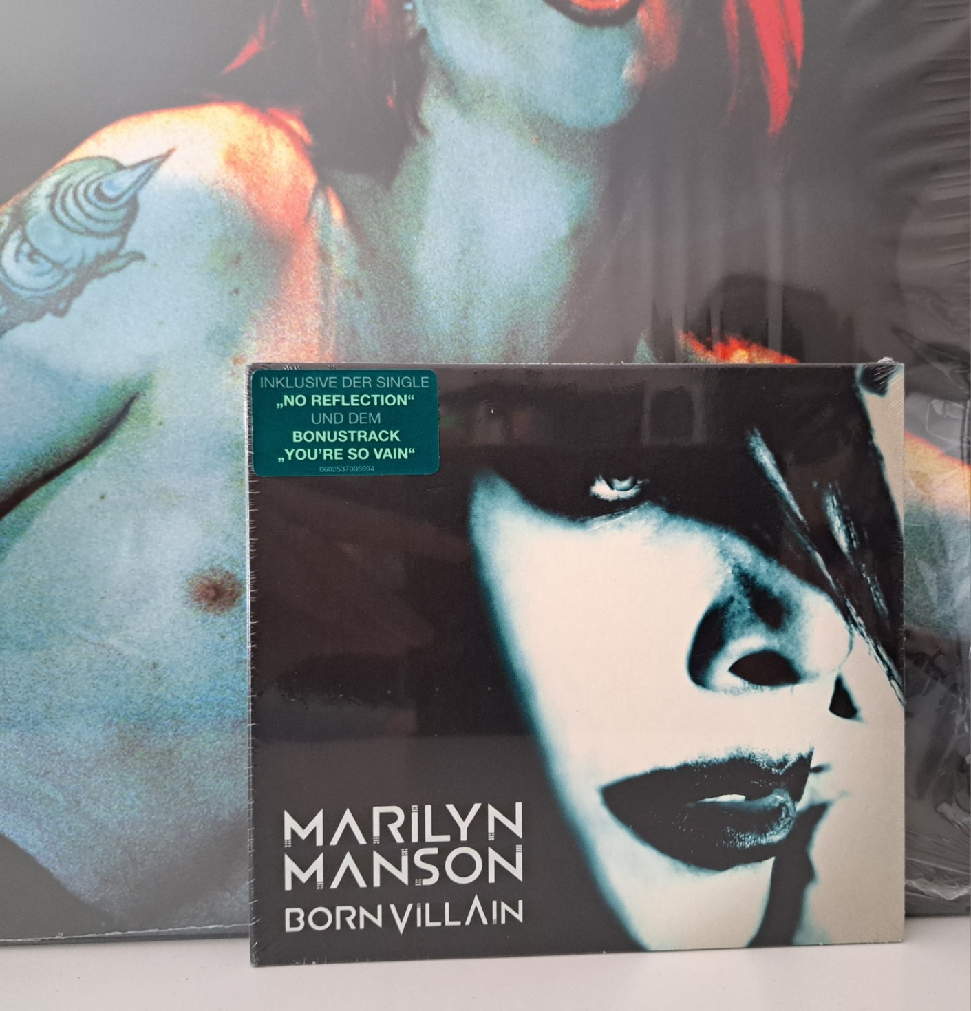 A picture of Marilyn Manson's Born Villain in front of a bigger Manson picture. Both are quite reflective as they're wrapped in plastic, but the CD has a sticker saying:  Inklusive der single "NO REFLECTION" und dem BONUSTRACK "YOU'RE SO VAIN"  While "You're So Vain" is announced as a bonus track, I challenge you to find an edition of this record without it (spoiler alert: as far as I know, there isn't one, and I've looked for it - at lot).