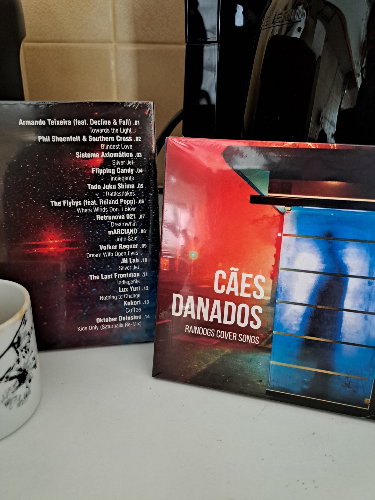 Two sealed copies of "Cães Danados - Raindogs Cover Songs" on CD, one showing the front and the other the back cover. They are in front of a coffee machine and next to a coffee cup.  The thirteenth track, from Kokori, is named "Coffee".