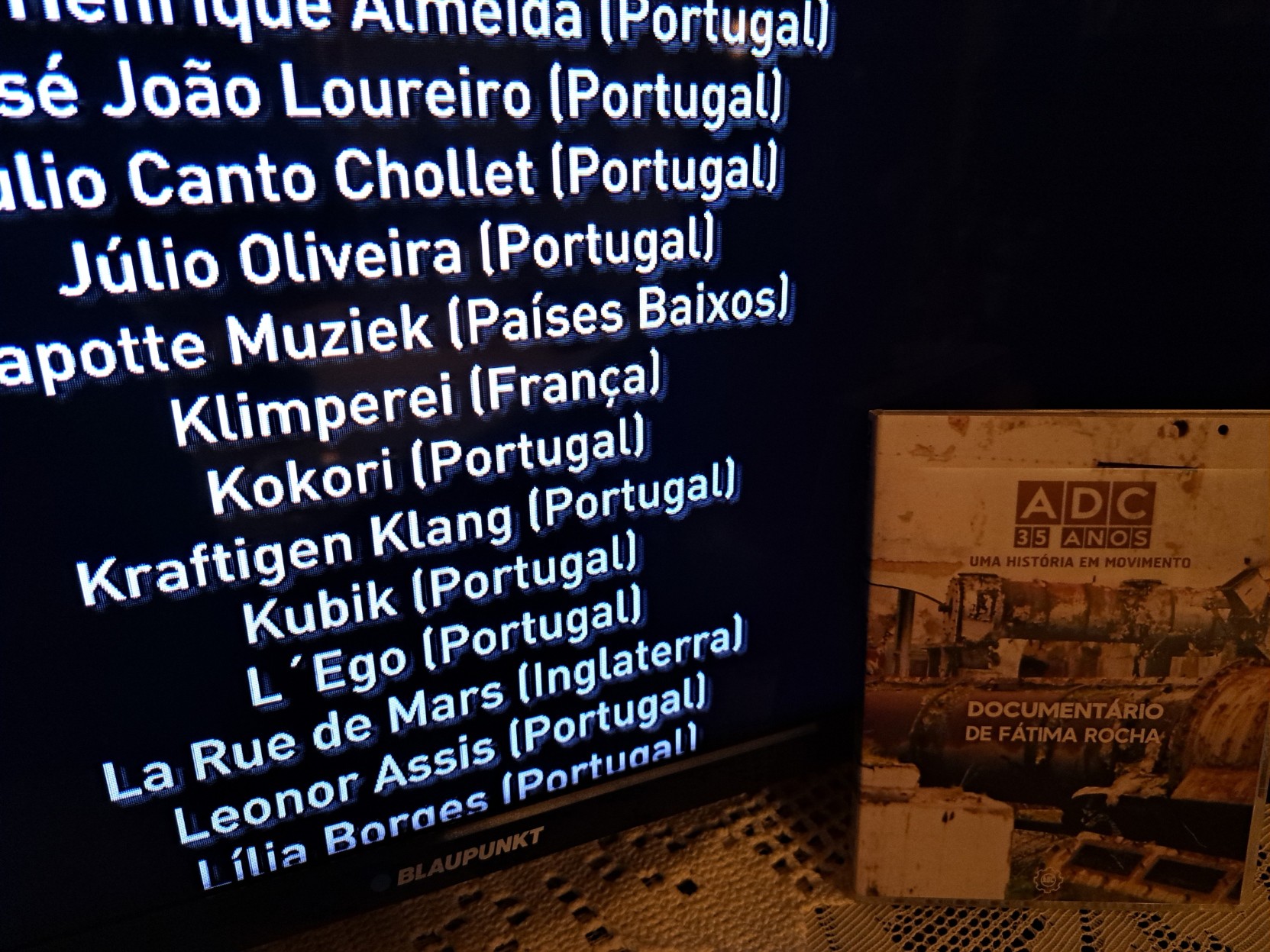Foto da caixa do documentário à frente de uma televisão onde o documentário está a passar - a mostrar uma lista de bandas relacionadas com a editora,vrais como os Kokori